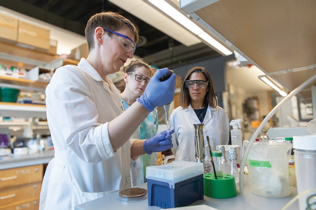 Kathryn Ramsey wears a labcoat and safety glasses, holding a pipette in her hand while two students in lab coats and safety glasses look on