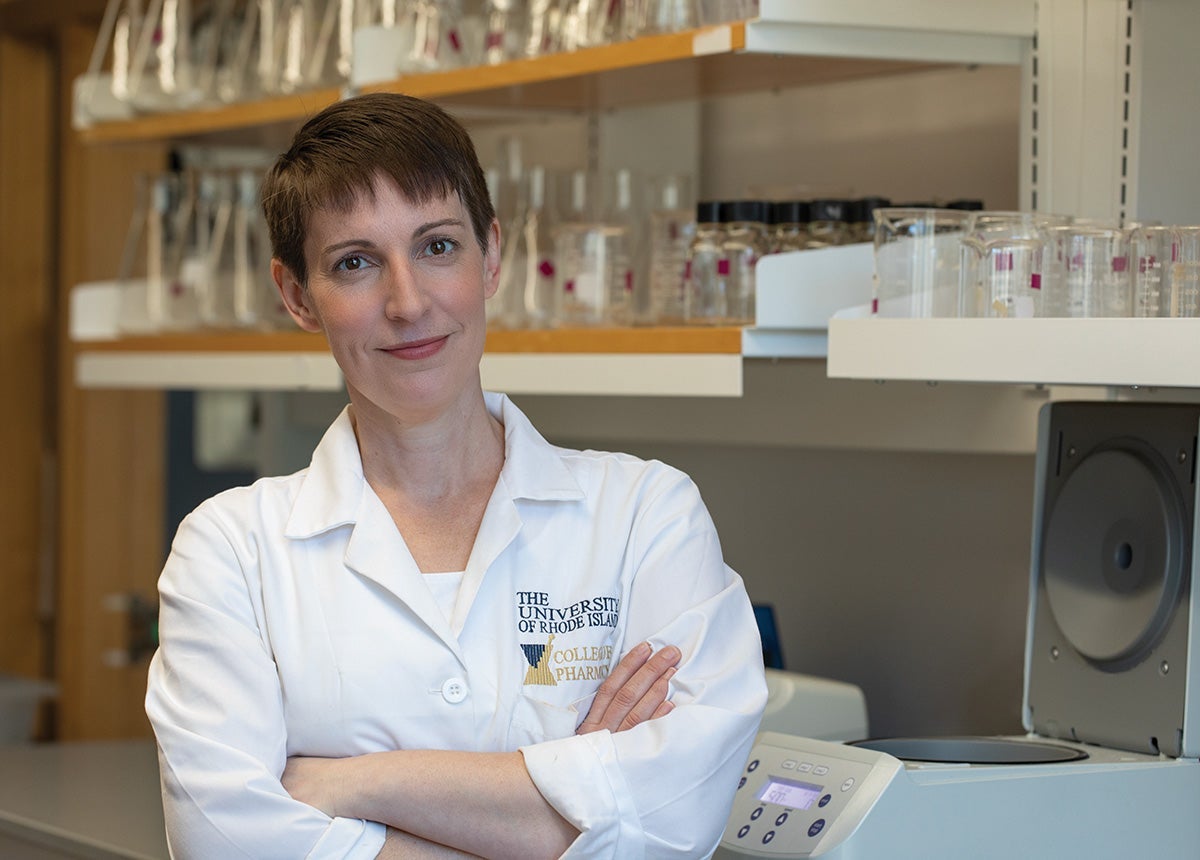 URI Professor Kathryn Ramsey stands in profile wearing white lab coat with arms crossed