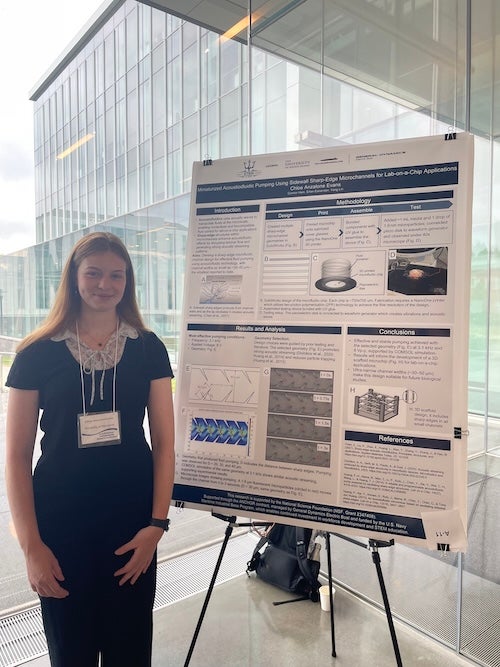 A young woman stands in front of a research poster in a well-lit glass-walled building.