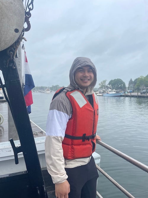 A young man wearing a hoodie and a life vest stands at the rail of a ship overlooking Narragansett Bay.
