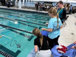 Two middle school students and one adult stand in front of a pool watching an aquatic robot built from PVC pipe navigate an obstacle course in the pool