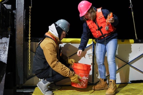 Two people in hard hats and life vests lift a bucket onto the deck of a ship.