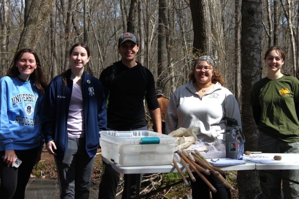 Five smiling students with field equipment stand among trees in the North Woods.