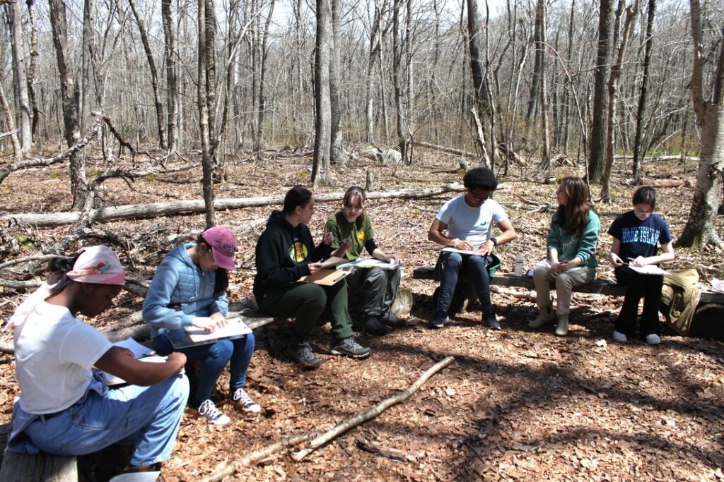 Students hold clipboards and notebooks while sitting on log benches.