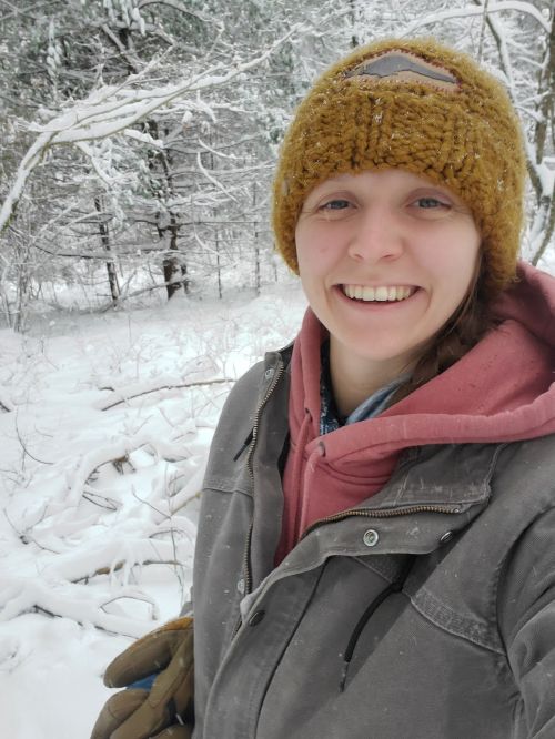 Laken Ganoe, wearing a gray winter coat, salmon sweatshirt, and mustard hat, smiling with snow and snow-covered trees in the background.
