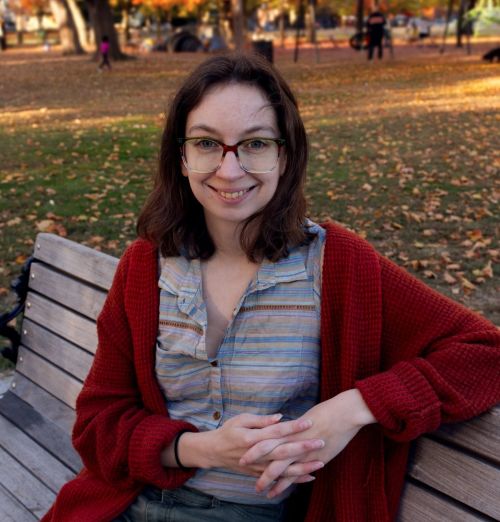 Abbie Lahmers, sitting on a wooden bench, wearing a red-orange cardigan, smiling, with grass and fallen leaves in the background.