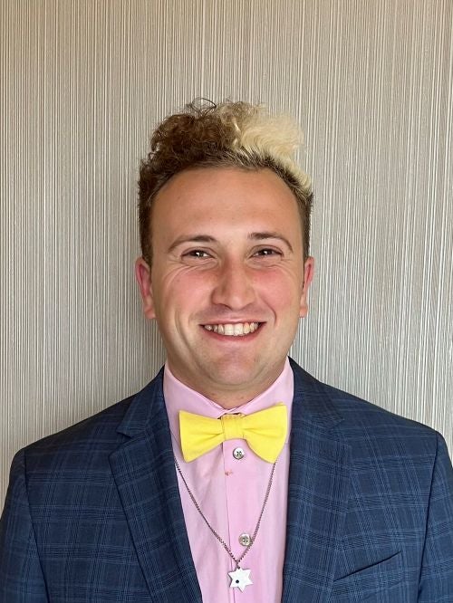 Ethan Brown, wearing a pink button-down shirt, blue sports jacket, and yellow bowtie, smiling against a white background.