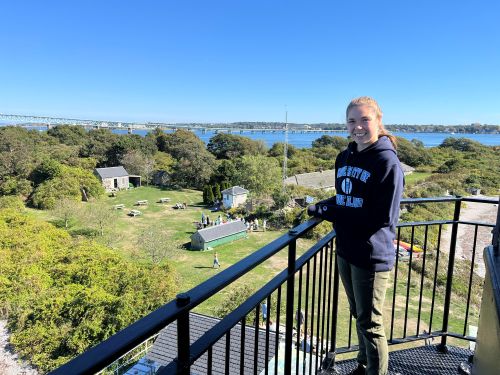 Sarah Heavren, standing on the balcony of Rose Island Lighthouse, overlooking green trees and bushes with ocean and blue sky in the background.