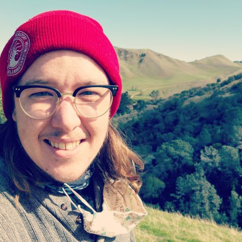 Shelby Rinehart, wearing a red beanie, smiling, with green grass, trees, mountains, and blue sky in the background.
