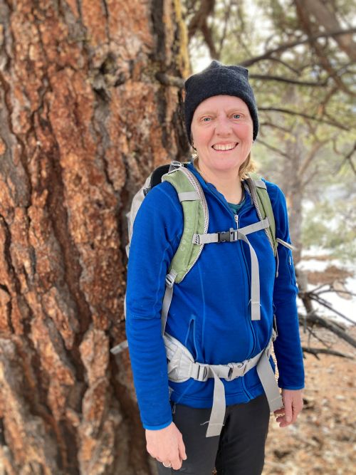 Nancy Karraker, wearing a blue coat, sage backpack, and black beanie, standing next to a large tree, smiling.