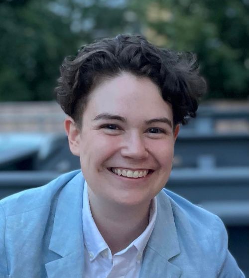 Mel Thibeault, wearing a light button down shirt and light gray sports jacket, smiling with trees in the background.