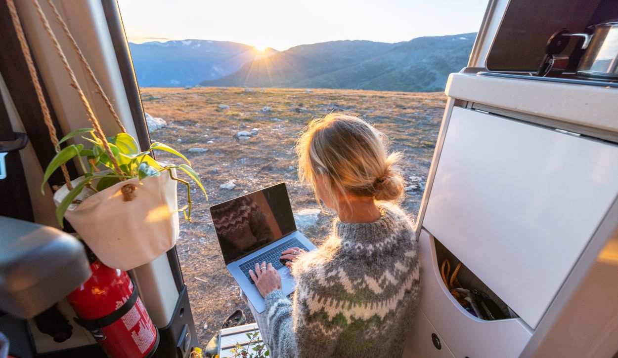 A blonde woman works remotely on her laptop from a van.
