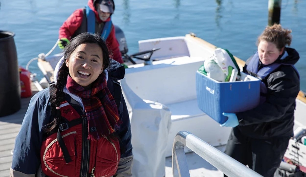The image depicts three people on a small boat docked beside a pier. In the foreground, a person with a red life jacket and a plaid scarf is smiling at the camera. The individual has long dark hair tied in a braid. To the right, another person, wearing blue gloves and a dark jacket, is carrying a blue storage bin filled with various items like bottles and containers. In the background, a third individual wearing a red jacket, a life vest, and a hat is adjusting a rope. The surroundings include calm blue water and part of a wooden pier.