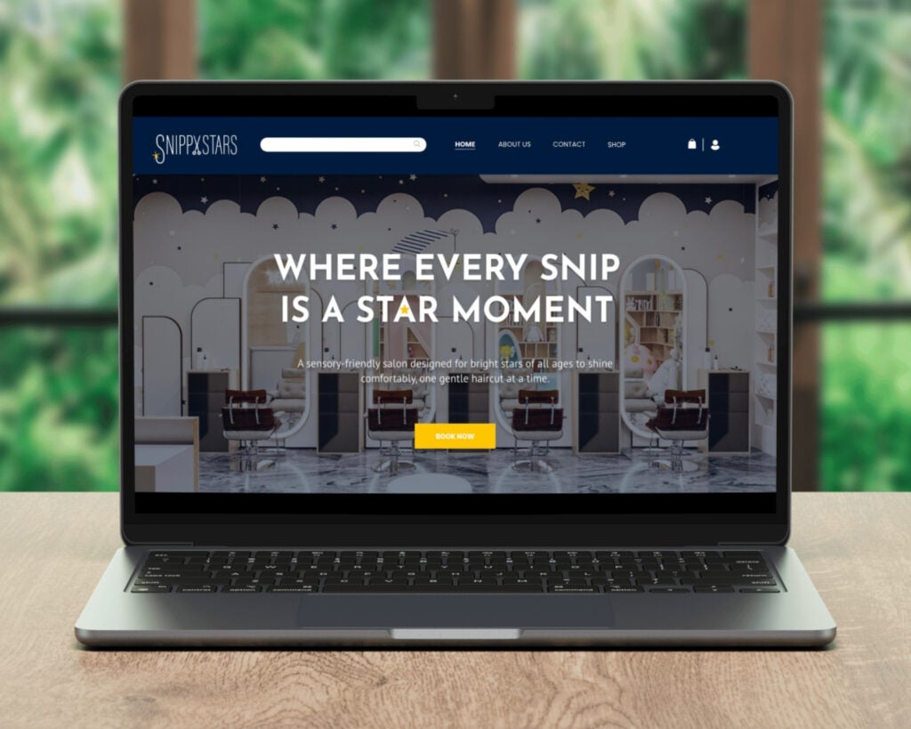 The image depicts a laptop on a wooden table with a blurred green garden background. The screen displays a website for Snipps & Stars, a salon with a child-friendly theme. The website features a main banner image of an interior salon space, characterized by a playful design with cloud and star motifs. The salon chairs are white and brown, facing mirrors designed in a circular shape. The navigation bar on the website includes links to Home, About Us, Contact, and Shop, along with search and shopping cart icons. A yellow "Book Now" button is prominently displayed below the main text.