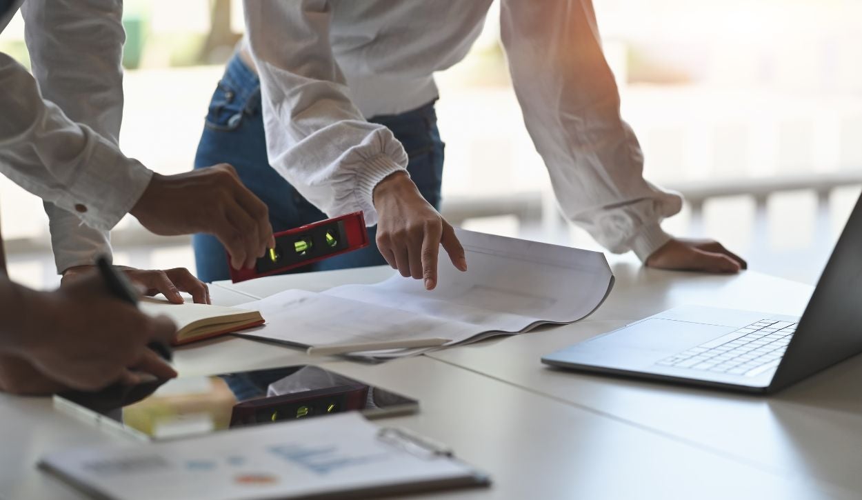 The image depicts a close-up view of a work desk where three people are collaborating. Two people are wearing white shirts and are visible from the waist down. One person is holding a red level tool, while the other is gesturing towards a large blueprint or paper spread out on the table. A third person, whose hand is partially visible, is holding a pen over a small open notebook. A laptop with a sleek design is situated on the right side of the table. The desk also contains a tablet and some documents. The scene is well-lit with natural light from the background.