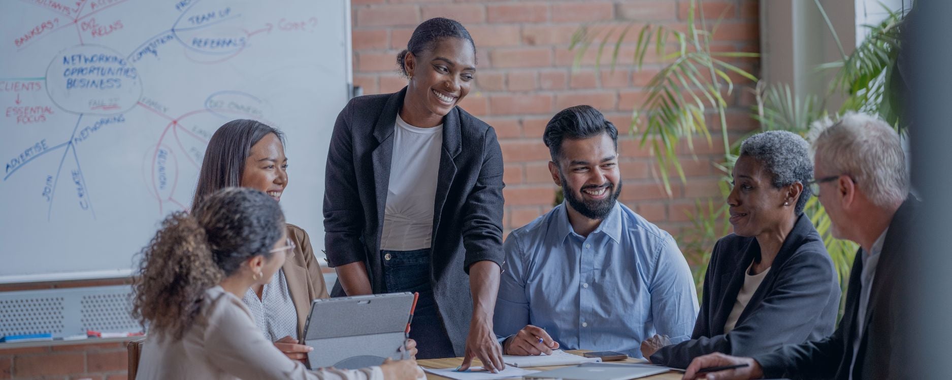 The image shows a diverse group of six people engaged in a meeting in a modern office setting. At the center, a person is standing and smiling, wearing a black blazer and a white shirt, appearing to lead the discussion. Around them, five others are seated around a table, listening and engaging with each other. The background features a brick wall and a large whiteboard filled with colorful handwritten notes and diagrams, partially visible, adding to the collaborative atmosphere. Nearby, a pot with green plants adds a touch of nature to the room. The lighting is bright and natural, coming from a window on the right.