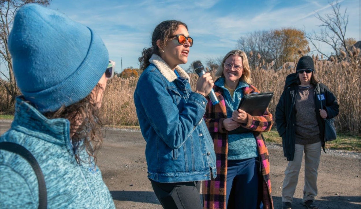 The image shows a group of four people outdoors on a sunny day. One person is speaking into a microphone and wearing a blue denim jacket with a white fleece collar and sunglasses. Another person, standing to their right, is smiling and holding a black folder, dressed in a colorful checkered coat, with glasses and blonde hair. A third person stands to the left, partially visible, wearing a blue knit hat and sunglasses. The fourth person is in the background holding a bag over their shoulder, wearing dark sunglasses, a black beanie, and a dark jacket. The setting is a rural area with dry reeds and grass in the background under a blue sky.