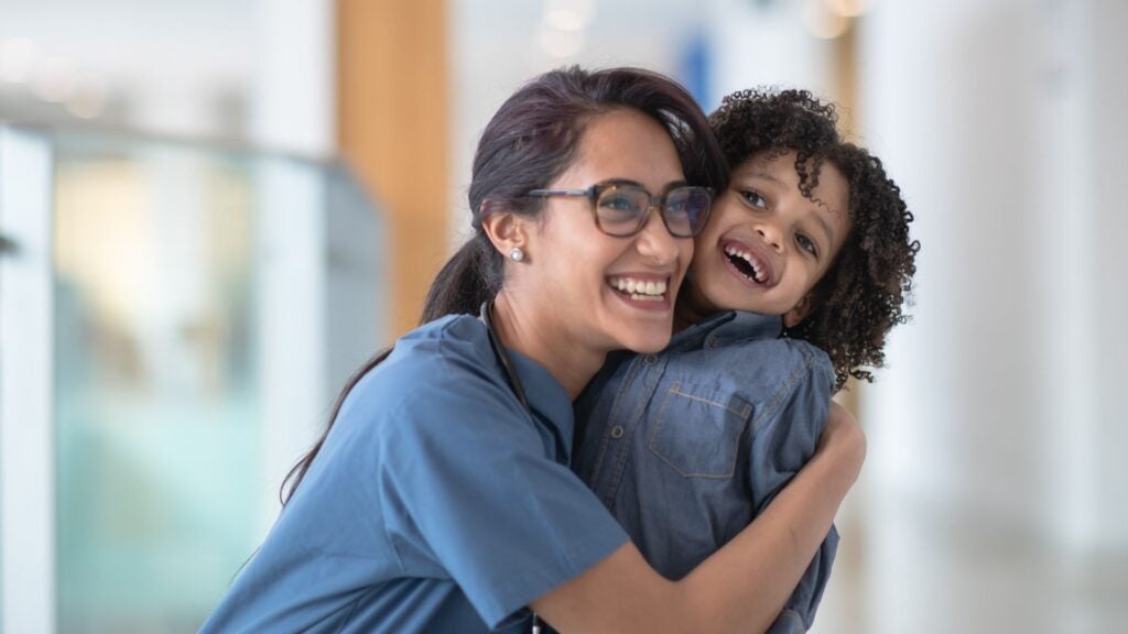 A woman in a blue medical uniform hugs a laughing child.
