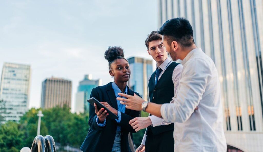 The image depicts three people engaged in conversation in an urban outdoor setting. On the left, a person with curly hair is holding a tablet and wearing a dark blazer over a light blue shirt. In the center, a person in a white shirt and dark vest is listening attentively. On the right, a person is gesturing with their hands, as if explaining something, and wearing a white shirt. The background includes modern office buildings with glass façades and a clear sky.