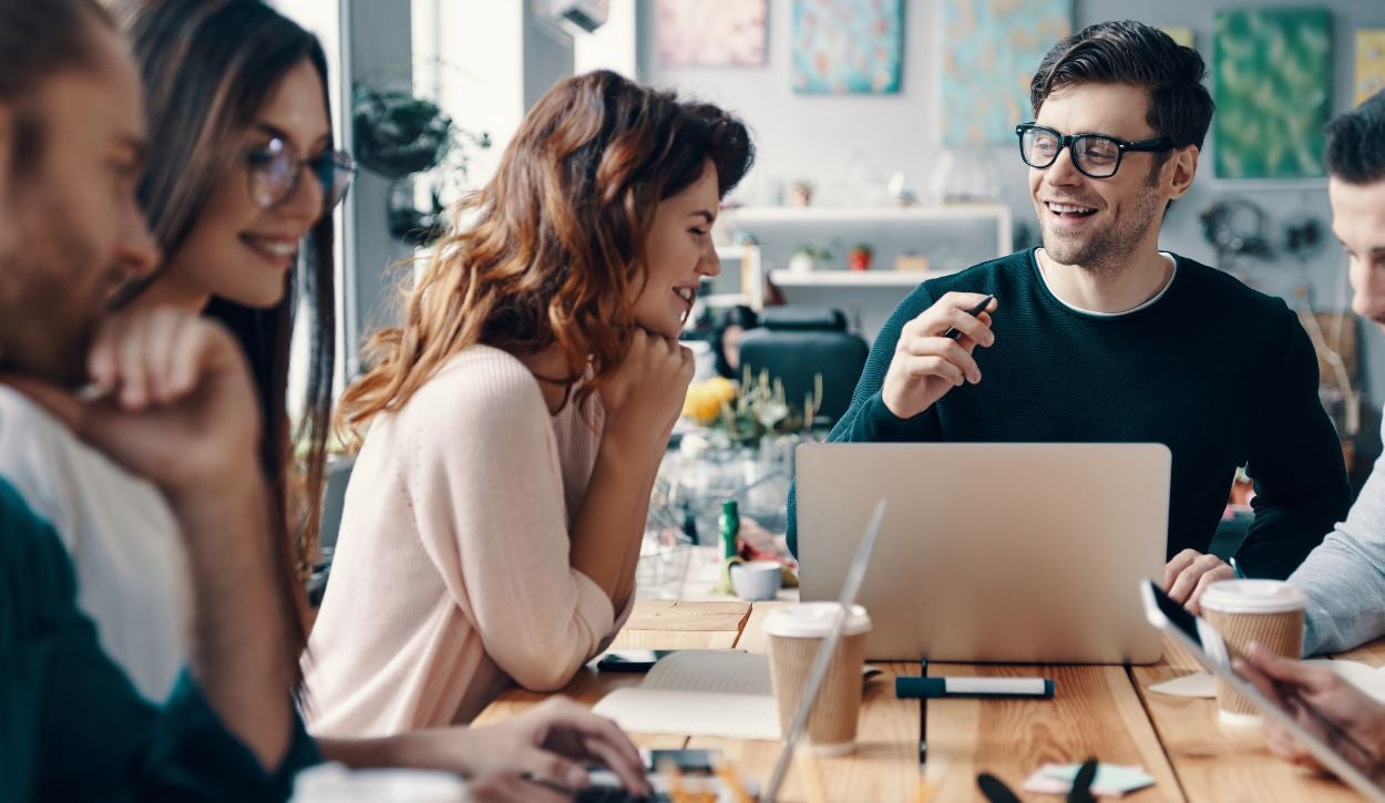 The image depicts a group of five people engaging in a discussion around a wooden table. The setting appears to be an informal meeting or brainstorming session. The individuals are casually dressed and appear to be talking and smiling, indicating a friendly and collaborative atmosphere. A laptop is open in front of the man at the center, who is holding a pen and appears to be speaking. Various items, such as notepads, coffee cups, and a smartphone, are scattered on the table. In the background, the room is decorated with abstract art on the walls and various decorative items on shelves.