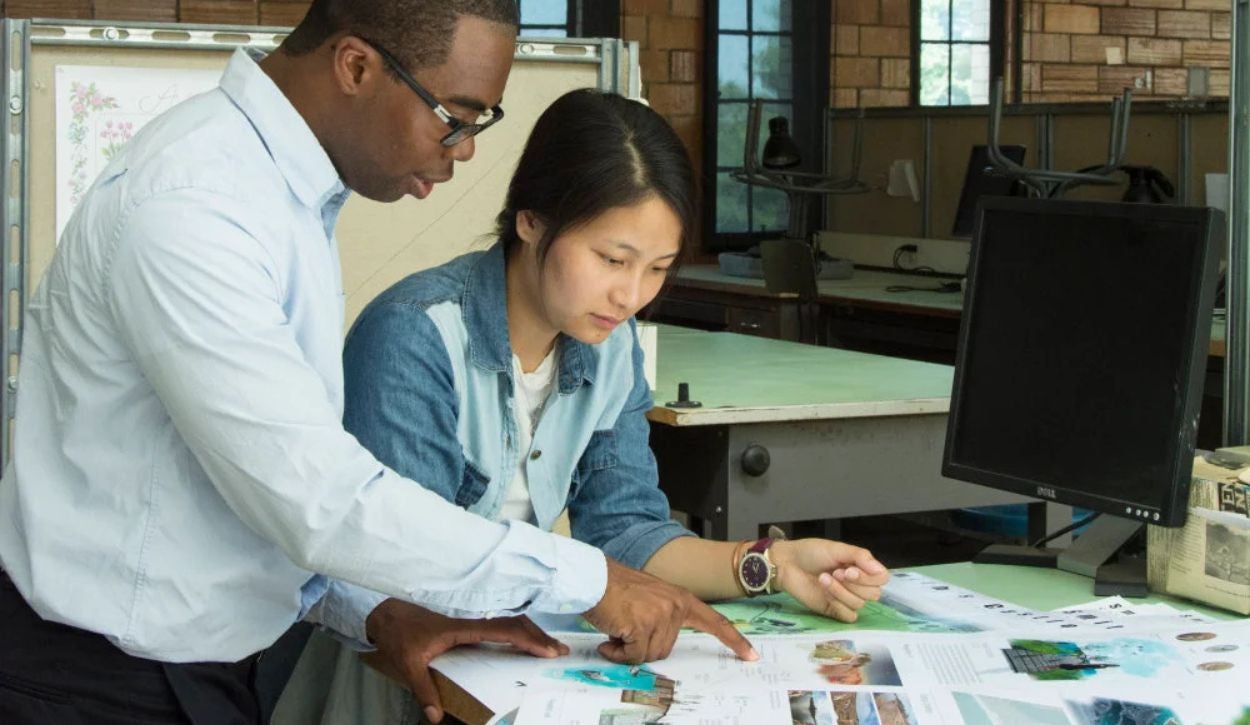 The image shows two people engaged in a discussion at a work desk in what appears to be a classroom or office setting. The person on the left is wearing a white shirt and glasses, pointing at documents spread on the table. The person on the right, wearing a denim jacket over a white shirt, is seated and looking attentively at the area the other person is pointing to. On the table are various papers with colorful images and text. Behind them, a computer monitor is visible, along with a background that includes large windows, an architectural drawing board, and several desk lamps.