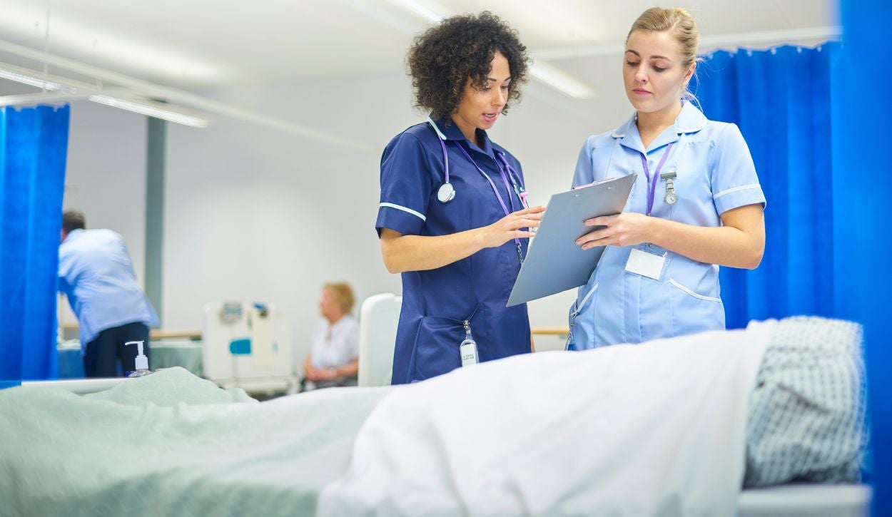 The image shows two healthcare professionals standing next to a patient's bed in a hospital setting. They are engaged in discussion, reviewing a document on a clipboard. One of the professionals is wearing a dark blue uniform and has a stethoscope around their neck, while the other wears a light blue uniform. Both uniforms have white trim. The room features blue privacy curtains and is brightly lit with a white ceiling. In the background, another healthcare worker and a patient are visible amidst hospital equipment.