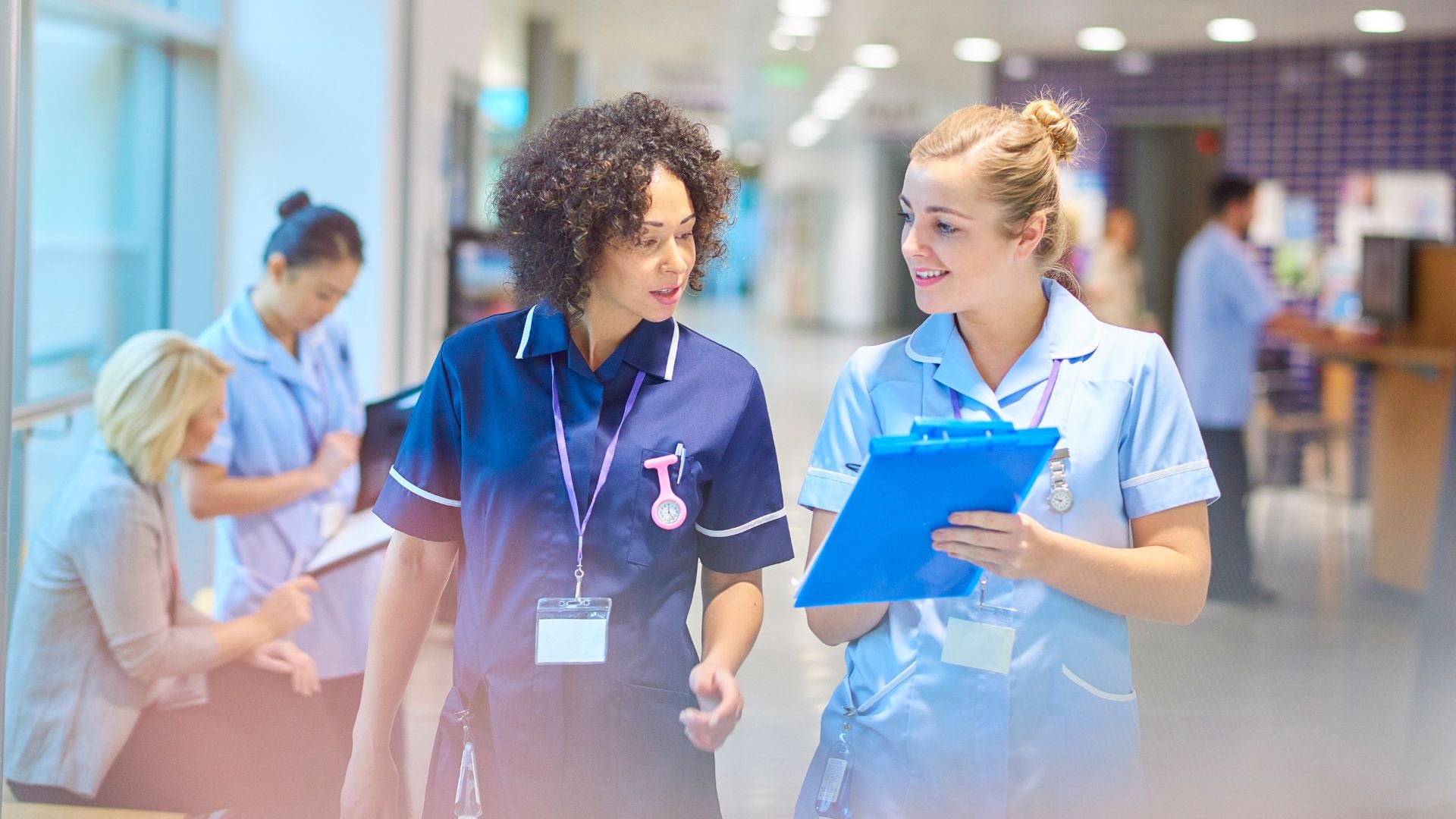 The image shows two nurses walking through a hospital corridor. The nurse on the left is wearing a dark blue uniform with a pink watch pinned to her chest, and she has curly hair. She is looking at the nurse on the right, who is wearing a light blue uniform and holding a blue clipboard. The nurse on the right has blonde hair tied up and is smiling. In the background, there are two additional nurses and a seated woman, all engaged in their work. The corridor is well-lit with overhead lights and blurred activity in the background, indicating a busy hospital environment.