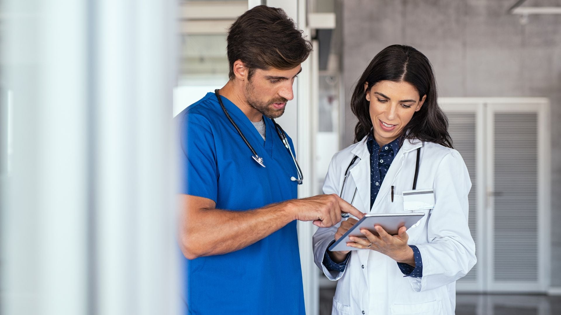 The image shows two healthcare professionals inside a medical facility. A male doctor on the left, wearing blue scrubs and a stethoscope around his neck, is pointing at a tablet held by a female doctor. The female doctor, on the right, is wearing a white lab coat over a dark shirt with small patterns, and she has a stethoscope as well. They are both focused on the screen of the tablet. The background is blurred, showing modern gray walls and part of a glass door.