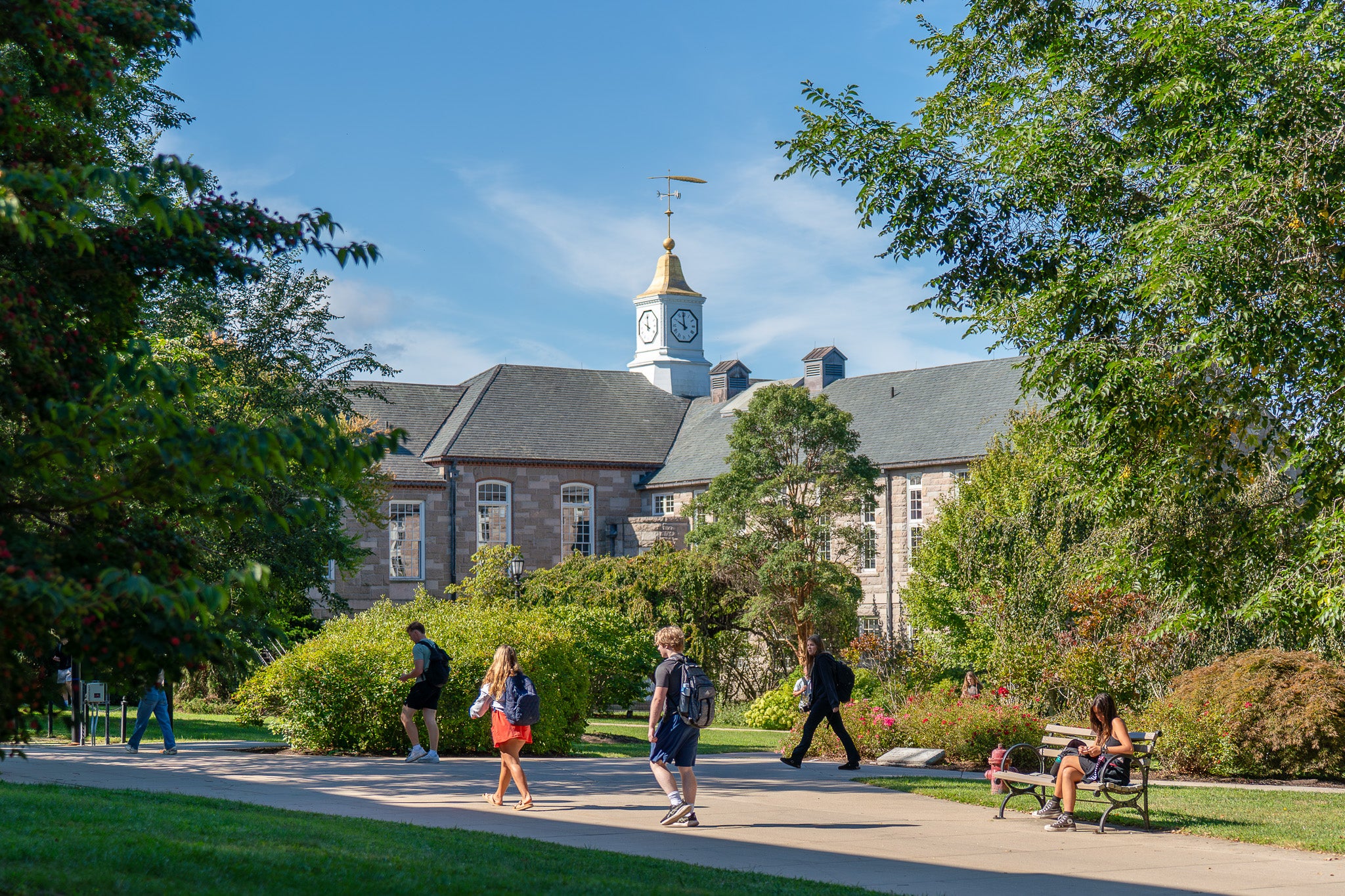 The image depicts a university campus scene with a building featuring a clock tower. The building is made of light-colored stone with a dark roof, featuring multiple arched windows. Above the building is a prominent clock tower with a golden cap and a weather vane. The foreground consists of a walkway with students casually walking, some carrying backpacks. On the right, a student sits on a bench amidst lush green foliage and vibrant landscaping, under a clear blue sky.