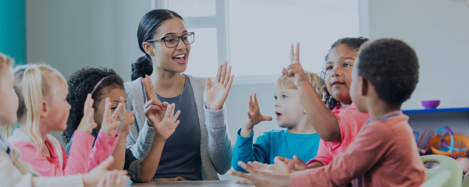 The image depicts a lively scene of a classroom with a teacher and several children participating in an interactive activity. The teacher, seated at the center, is wearing glasses and a gray sweater over a blue shirt, smiling and gesturing with her hands raised. Around her, five children are also engaged, imitating her gestures. The children, both boys and girls, are seated at a table and appear to be singing or counting along. The background shows a brightly lit room with large windows and colorful shelves holding various toys. The atmosphere is joyful and animated, indicating a playful learning environment.