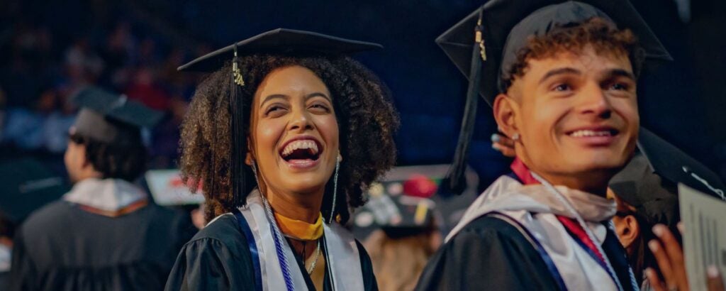 Two joyful graduates in caps and gowns at a ceremony, laughing and smiling.