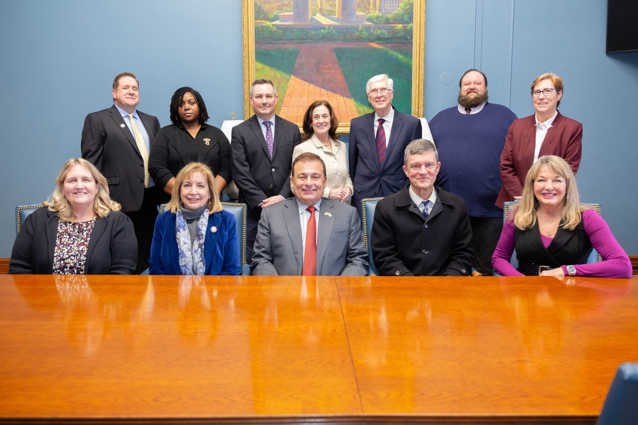 A group of MPA faculty members, URI leadership, and RI State Representatives pose in a formal setting, seated and standing in front of a wooden table, with a large painting behind them.