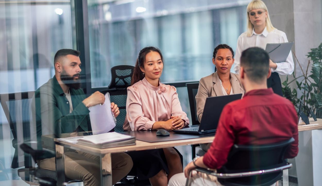 The image shows a modern office setting with four individuals seated at a glass conference table and one person standing. The person in the foreground, facing away from the camera, is wearing a red shirt and is engaging with the other seated individuals. To the left, a bearded man in a dark green jacket holds papers. In the center, a woman with a light pink blouse sits with her hands resting on the table beside a laptop. To her right, a woman in a beige blazer is also seated attentively. A standing person in the background, wearing a white shirt and glasses, holds a clipboard. The room features floor-to-ceiling windows with a view of adjacent buildings, and several potted plants are visible.