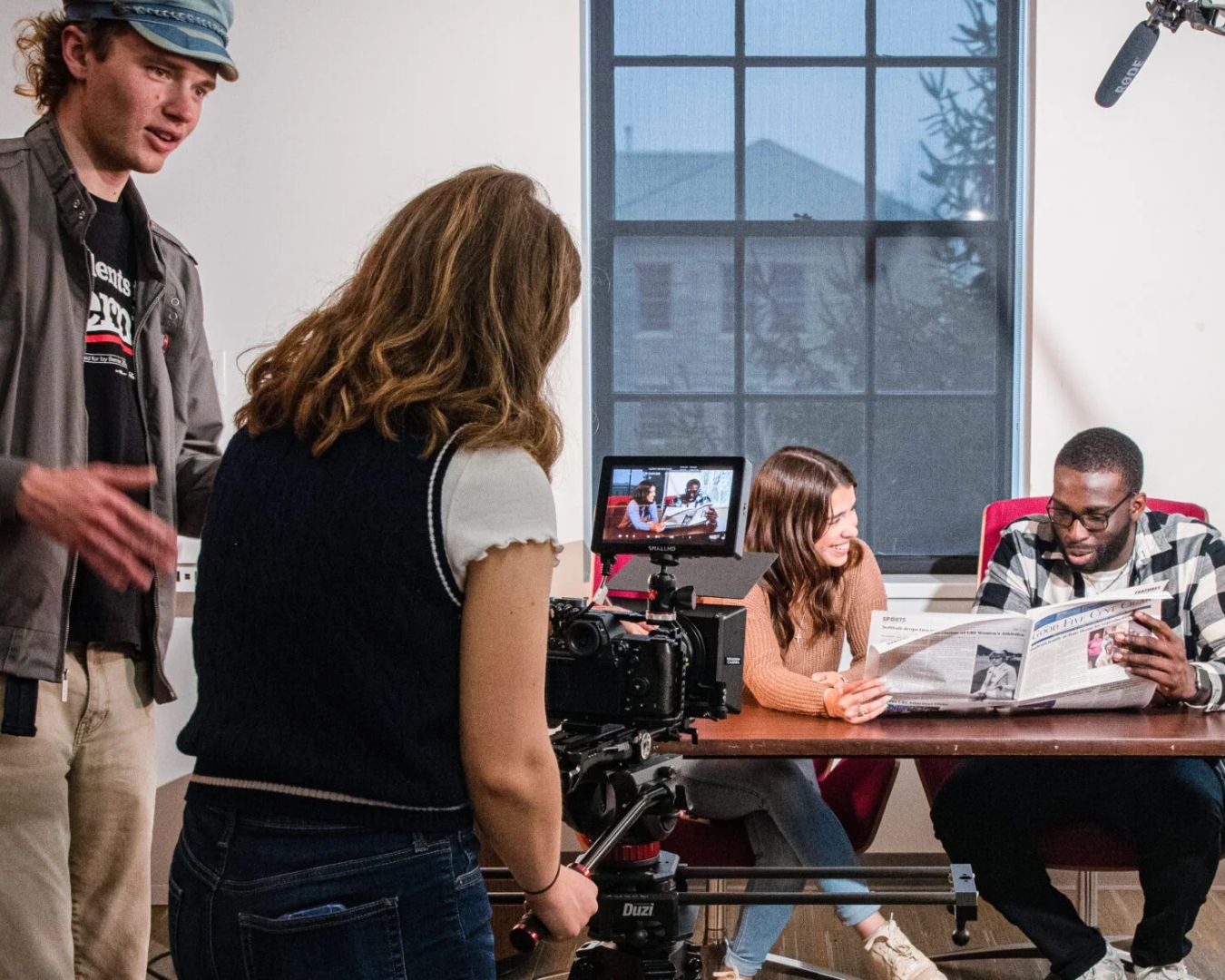 The image depicts a scene in a brightly lit room where a video shoot is taking place. In the foreground, a woman with long hair, wearing a navy blue vest and white shirt, operates a professional camera mounted on a tripod. The camera screen displays a live view of the shot, showing two people sitting at a table. To the right, a woman with long dark hair, dressed in a brown sweater, smiles and looks towards her companion, a man in a plaid shirt, who is holding and reading a newspaper. The background features a large window with a grid design, through which a silhouette of a tree is visible. On the left side, another person, wearing a cap and light jacket, gestures as if giving direction. A microphone is suspended above the setup, indicating a formal recording session.