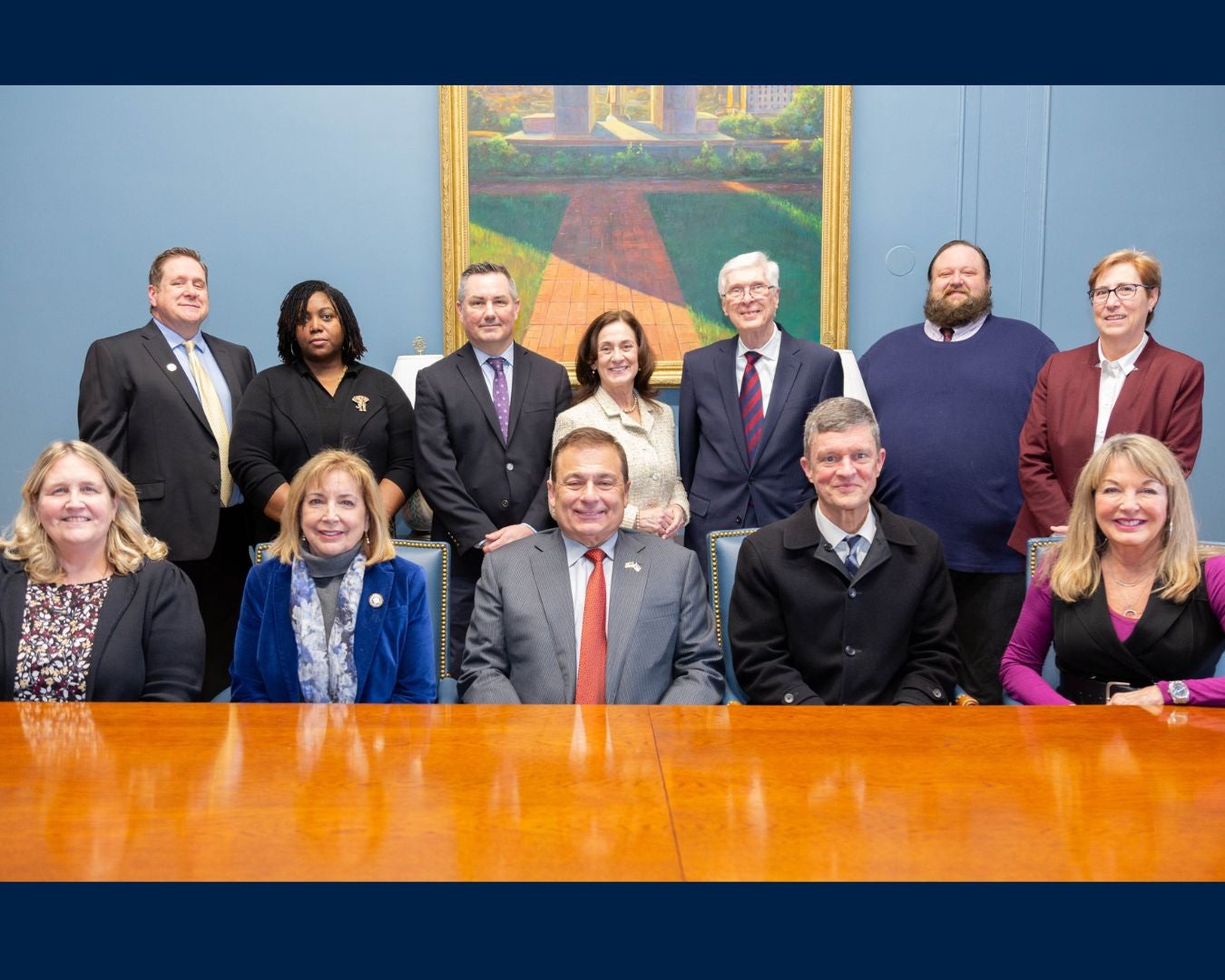 A group of MPA faculty members, URI leadership, and RI State Representatives pose in a formal setting, seated and standing in front of a wooden table, with a large painting behind them.