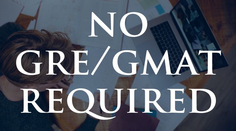 The image contains the large text "NO GRE/GMAT" over a blurred background that shows a person working at a desk with an open laptop, books, and papers scattered around. The text indicates that GRE or GMAT test scores are not required, likely in the context of academic programs or admissions.