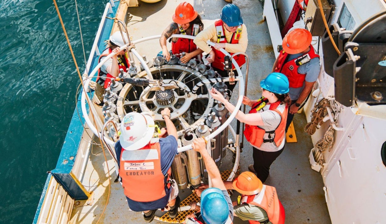 The image shows a group of seven individuals on the deck of a boat gathered around a complex cylindrical scientific instrument. The instrument is positioned close to the ship's railing over the water, indicating a marine research setting. All individuals are wearing orange life vests and various colored hard hats, with safety equipment visible. The device has multiple cylindrical sampling containers arranged in a circular structure. There are ropes and wires connecting different parts, indicating a deployment or retrieval operation. The boat's surface is metallic gray, showing signs of use, and there is a notable contrast against the deep blue water.