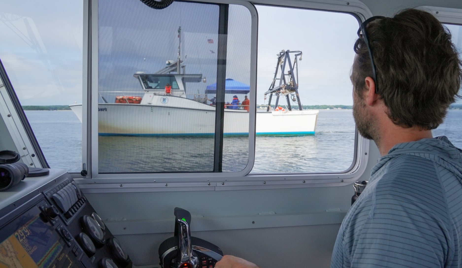 The image depicts the interior of a boat's cabin where a man is observing an adjacent ship through a large window. The man is seated, seen from behind, and appears to be focusing on the vessel visible outside. The cabin is equipped with various navigation instruments and controls. The outside vessel is a medium-sized fishing boat, visible through the window, featuring a blue trim, a white hull, and orange life rings. A group of people wearing bright clothing is seen on the deck. The sky is overcast, and the water is calm, creating a serene maritime environment.