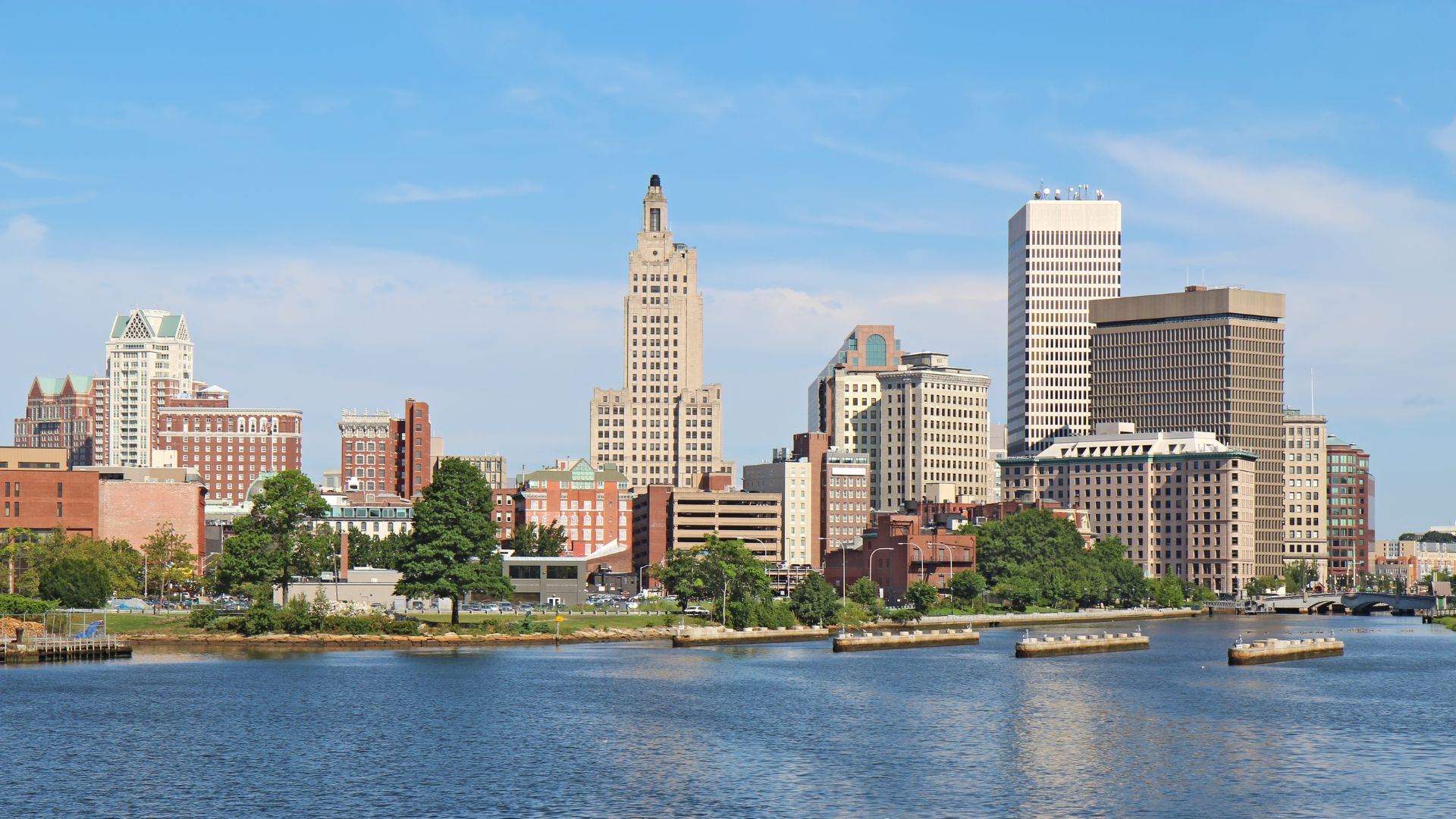 The image depicts the Providence city skyline with an array of high-rise buildings against a clear blue sky. In the foreground, a calm body of water reflects the structures and the greenery along the shoreline. The skyline features a variety of architectural styles, with a prominent tall, beige building with a unique tower-like top as a central focal point. Surrounding this are buildings with diverse facades, ranging from brick red to light gray, showcasing both modern and historic architecture. A few trees are visible near the water, adding a touch of nature to the urban scene.