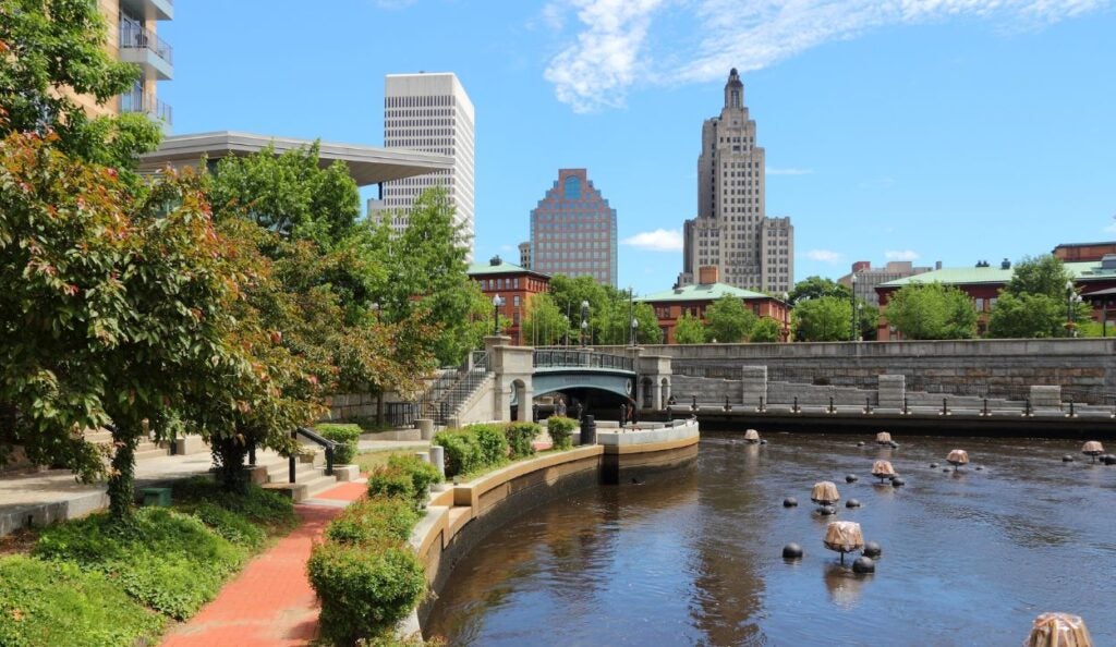 The image depicts a serene scene of Providence, Rhode Island featuring a river winding through the city with a combination of modern and historic architecture. On the left, a paved walkway bordered by lush greenery and red brick path runs alongside the river, shaded by trees with dense foliage. The river is calm, with evenly spaced stone structures partially submerged in the water. In the background, a small arched bridge crosses the river, leading to a series of buildings. Several skyscrapers dominate the skyline, including a tall, distinct building with an art-deco design featuring intricate patterns. The sky is bright blue, scattered with wispy clouds, suggesting a clear, sunny day.