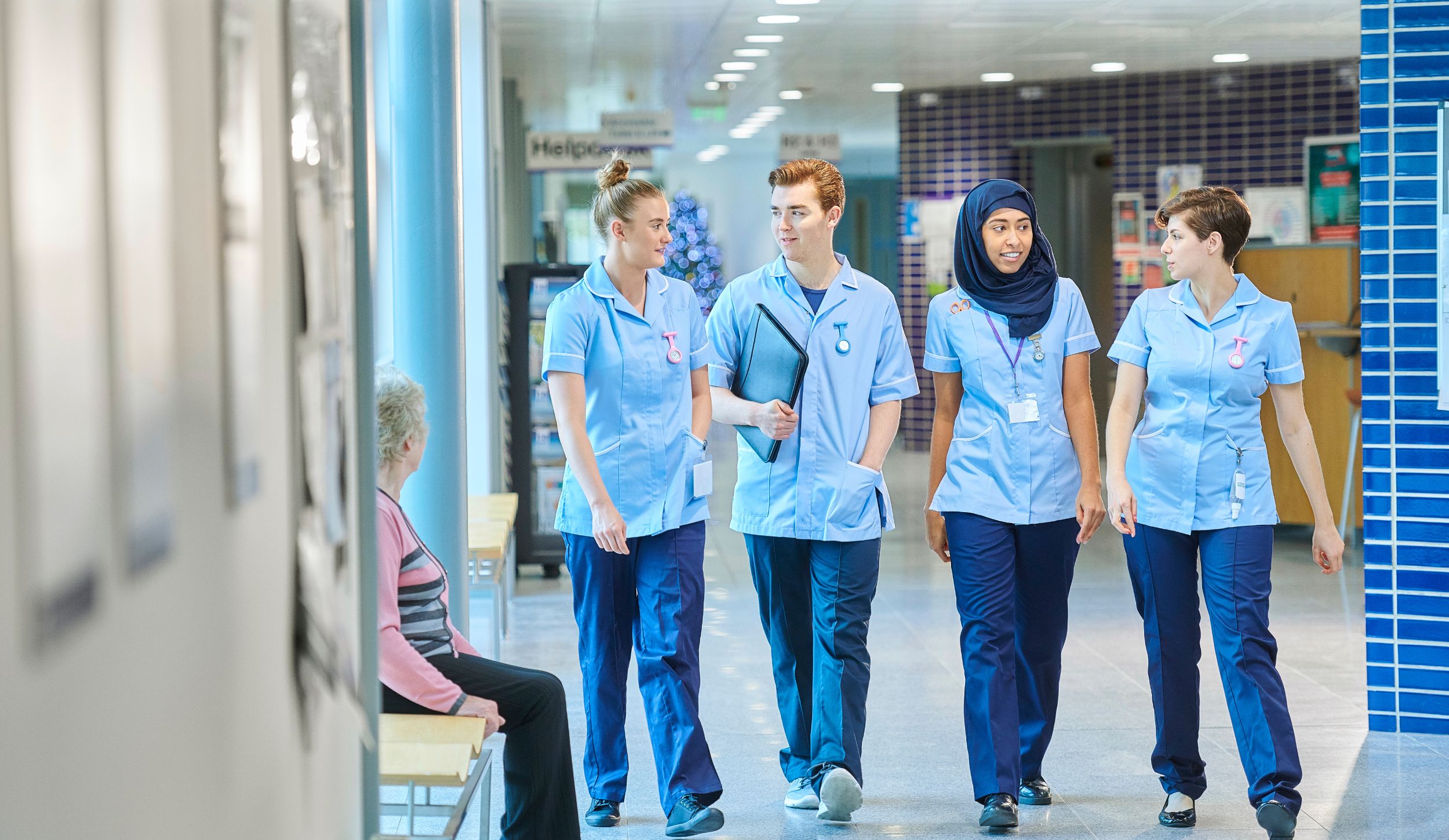 The image depicts a group of four healthcare professionals, possibly nurses, walking down a brightly lit hospital corridor. They are wearing matching light blue uniforms with navy trousers, and each carries a pen or ID clipped to their top. Their attire and badges suggest they are on duty. Behind them, there are various signs and blurred figures indicating a bustling medical environment. The corridor has a modern and clean design, with polished floors and bright overhead lighting. To the left, an elderly person with short, gray hair wearing a pink and black top sits on a bench, looking towards the professionals.