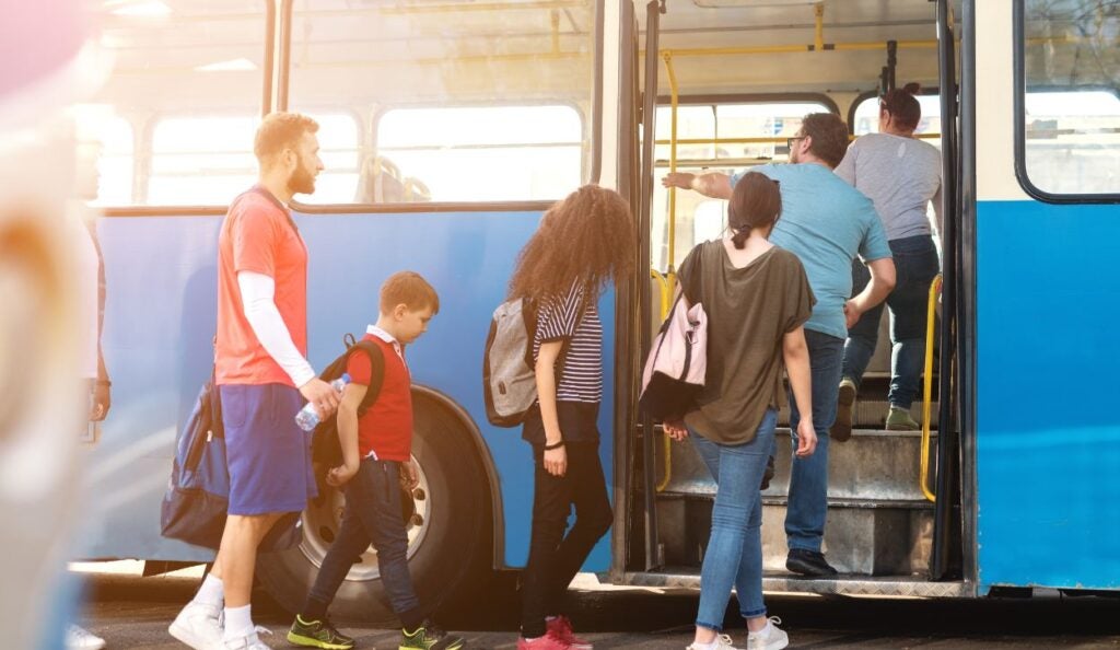 The image shows a group of people boarding a blue bus. A man in a red shirt and blue shorts is leading a young boy in a red top with a backpack. Behind them, a girl with long curly hair and a striped shirt carries a backpack while stepping onto the bus. Another person wearing a green shirt follows. The bus door is open, and several people are already stepping up into the vehicle. The bus interior is visible, with yellow handrails. The scene is brightly lit by sunlight.