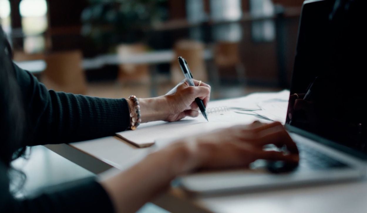 a person is writing on A piece of paper while sitting at a table with a laptop