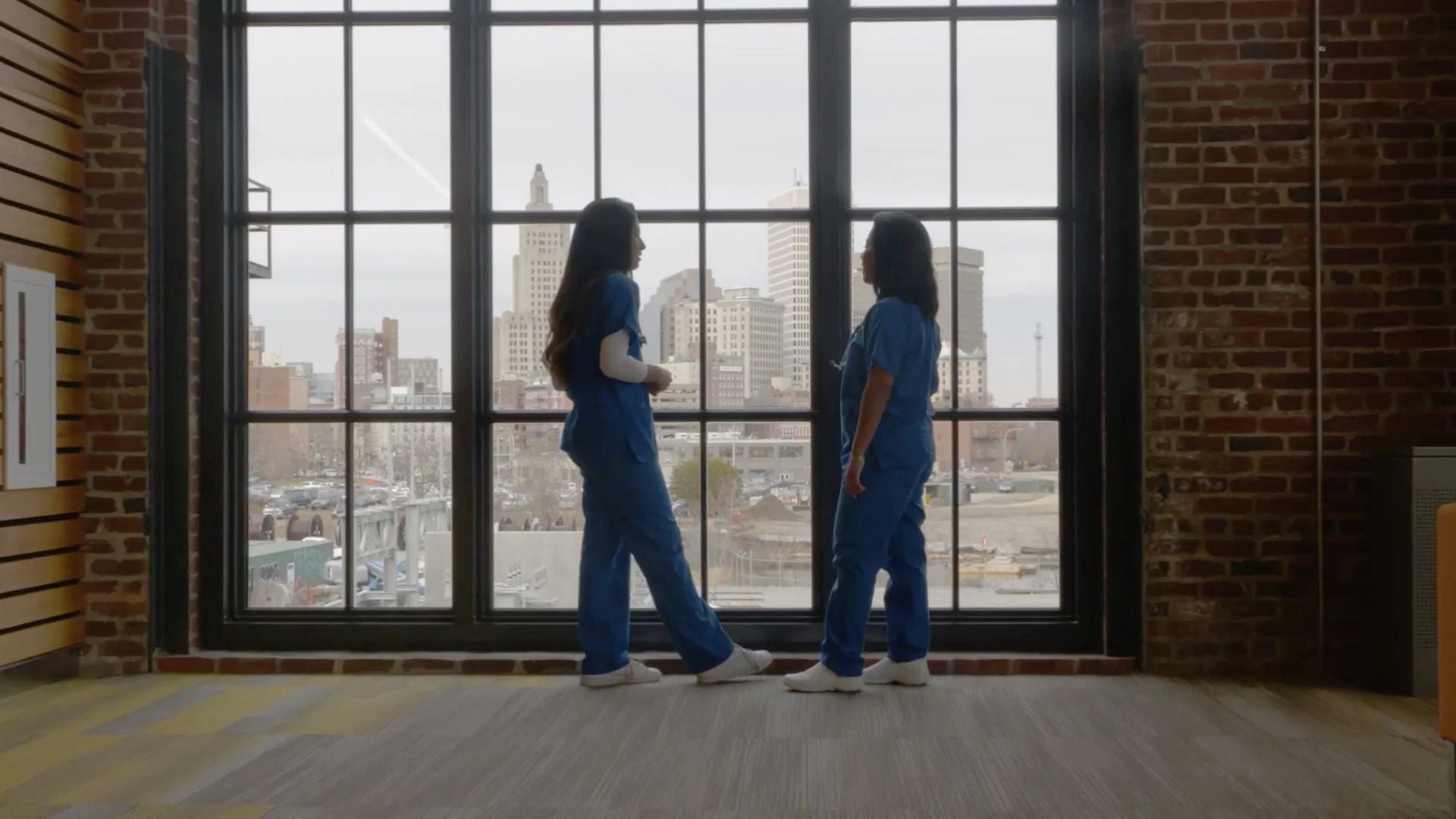 The image shows two people in blue scrubs standing in front of a large window with a grid design. They appear to be engaged in conversation and are looking out over a cityscape. The window reveals several tall buildings under a cloudy sky, with the city scene including a mix of modern and older architecture. The interior space features exposed brick walls and a floor with a subtle pattern. The lighting suggests it's daytime and enhances the natural tones of the environment.