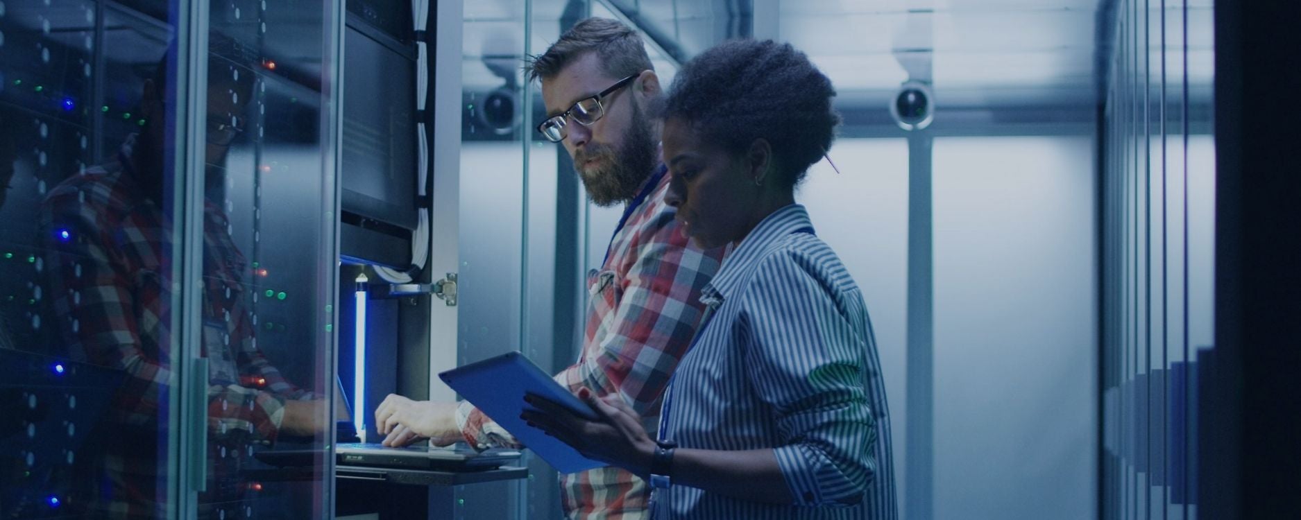The image depicts two people in a server room. The person on the left, wearing glasses and a red plaid shirt, is working on a laptop placed on a rack. They appear focused on the screen. The person on the right, wearing a blue and white striped shirt, holds a tablet and also appears engaged with their task. The server room is filled with tall racks housing electronic equipment, with several LED lights visible, casting a blue glow. The environment is sleek and modern, suggesting a high-tech setting.
