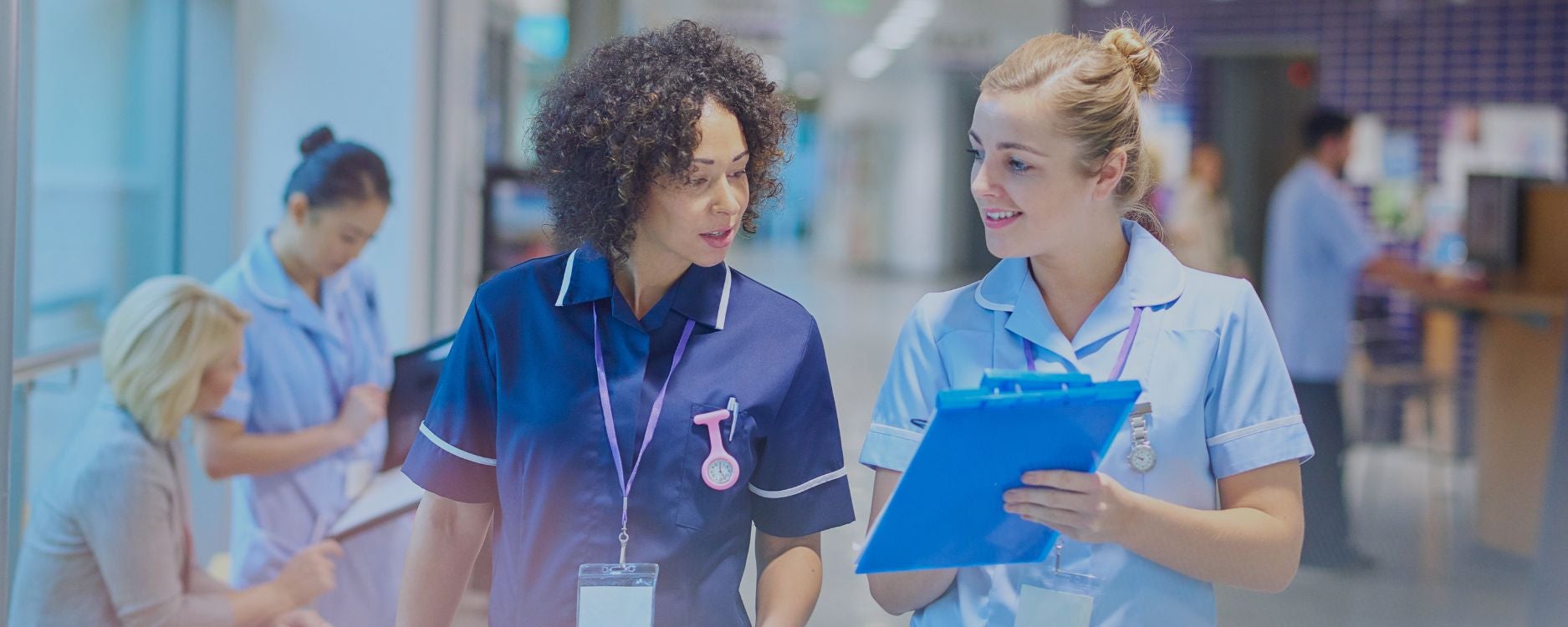 The image shows two nurses in a hospital setting engaged in conversation. One nurse, on the left, wears a navy blue uniform with white trim and a lanyard, and has curly dark hair. The other nurse, on the right, wears a light blue uniform with similar white trim, has her blonde hair tied back, and is holding a blue clipboard. In the blurred background, two more healthcare workers, also in light blue uniforms, are visible. The setting suggests a hospital corridor with glass walls and bright lighting.