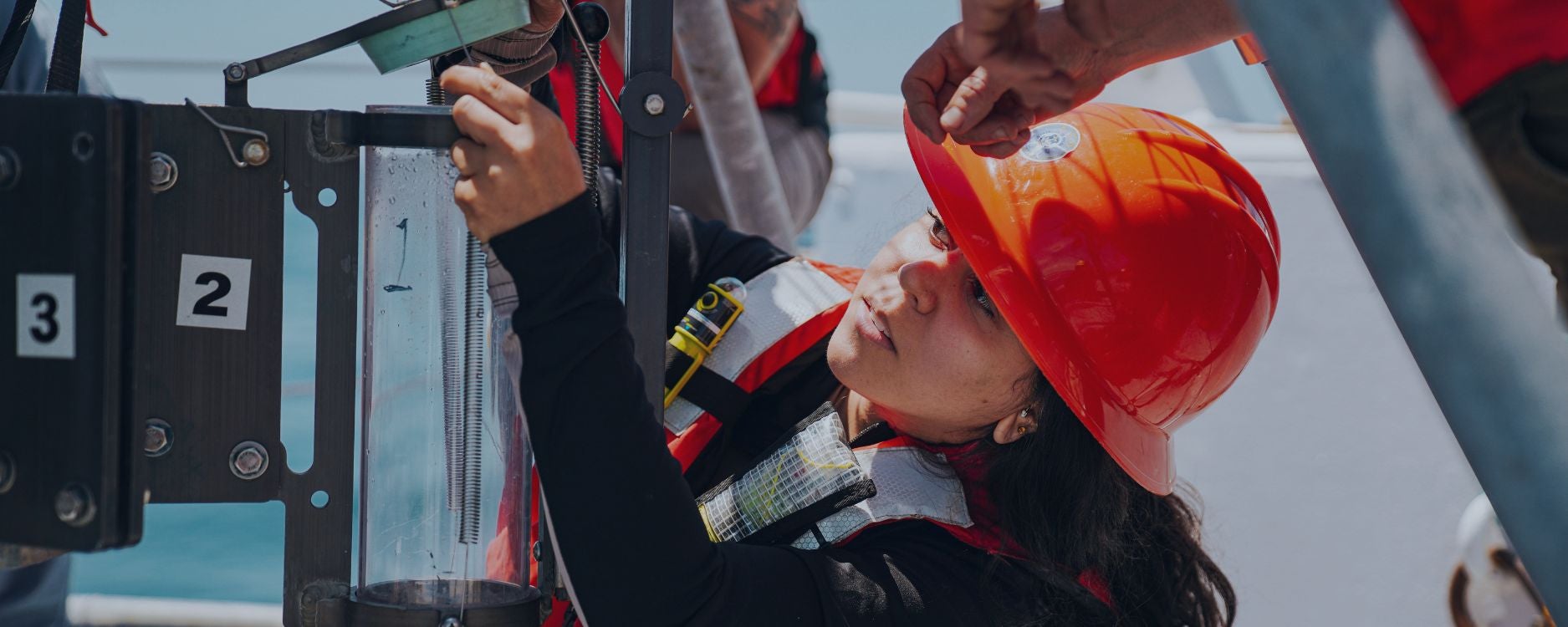 The image shows a person wearing a bright orange hard hat and a safety vest working on a piece of equipment. This individual is focused on adjusting a transparent vertical tube labeled with the number 