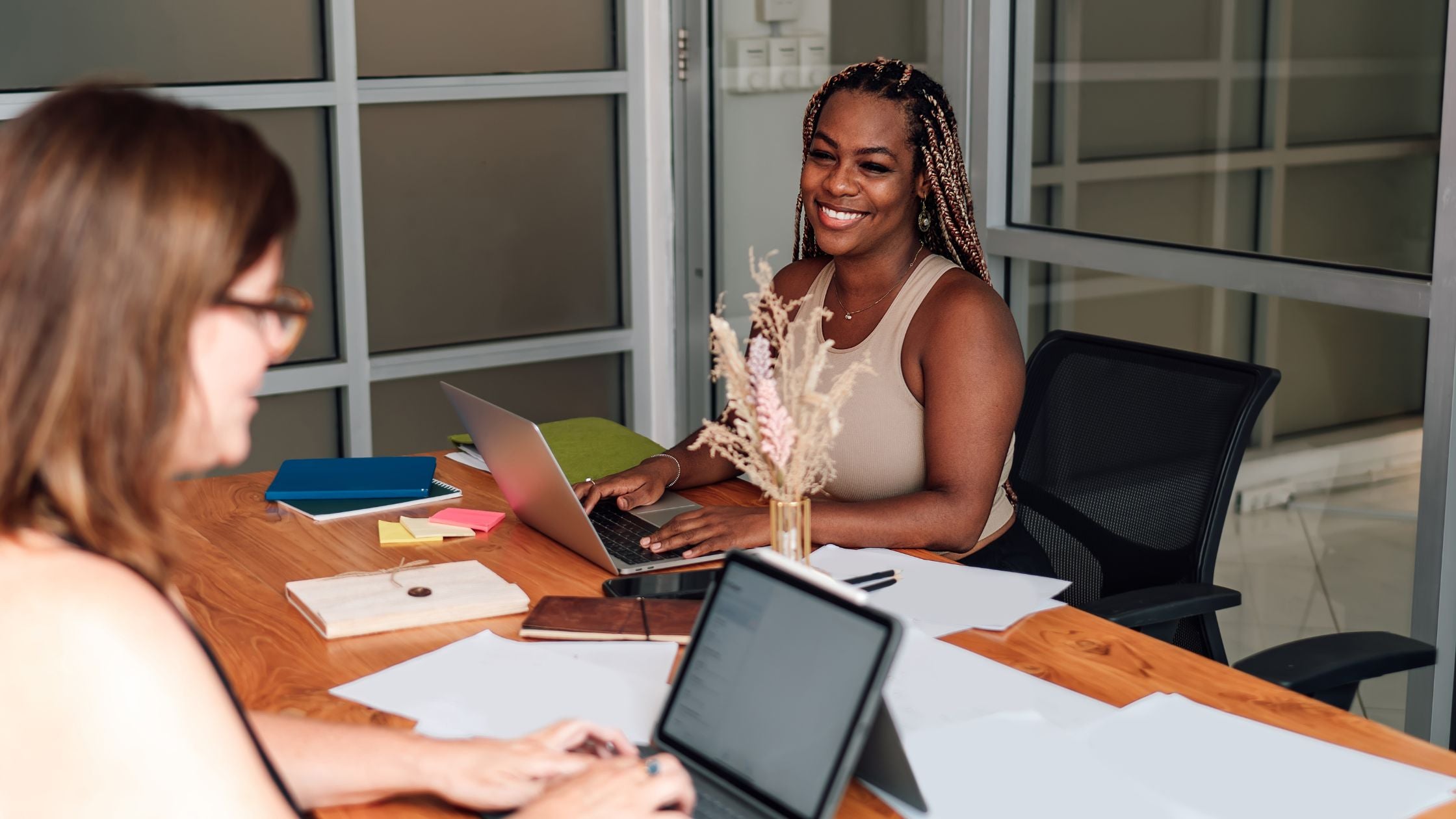 Two women smile and work together at a shared desk in a glass-walled office, with laptops, notebooks, and colorful sticky notes visible between them.