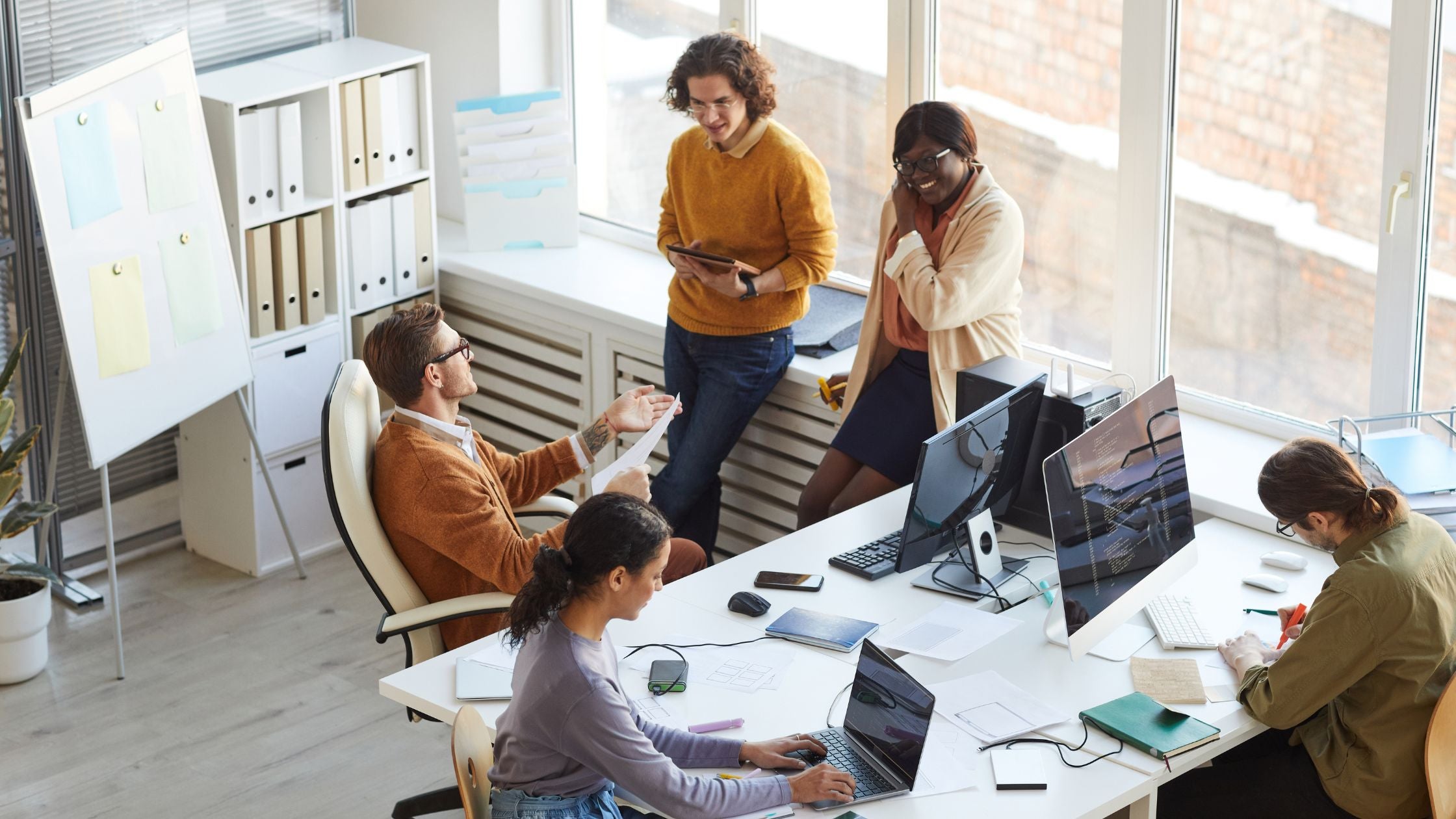 An aerial view of a diverse team working across a shared open-plan workspace, with some seated at computers, others standing and sharing documents in a bright, modern office.