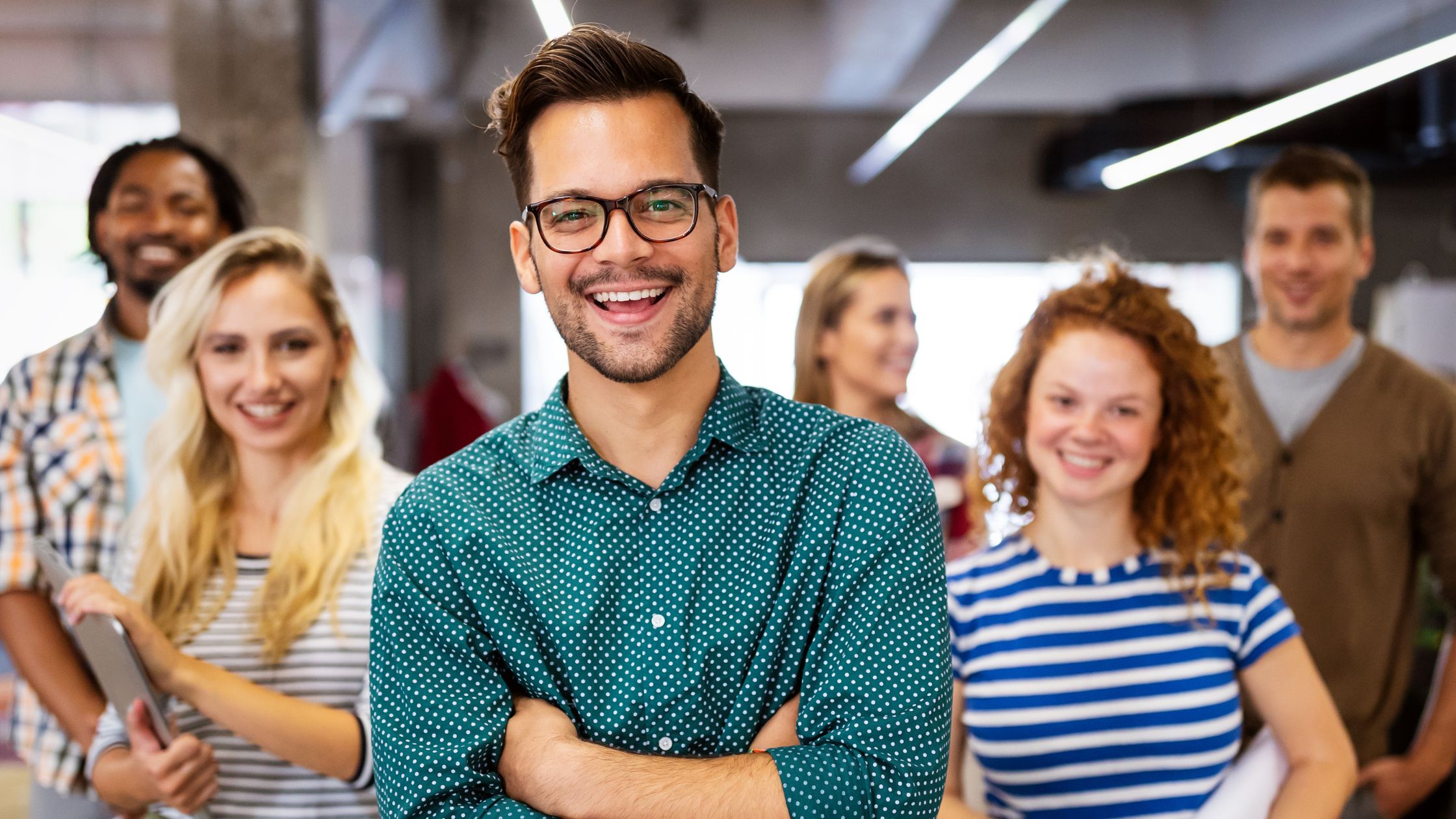 A young professional with glasses stands confidently with arms crossed and a wide smile, with a diverse group of smiling colleagues visible behind him.